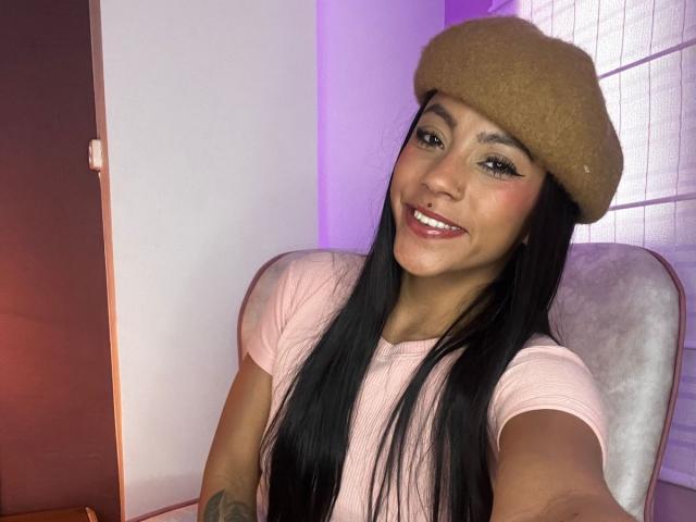 Amberv with long black hair, brown beret, pink t-shirt, and smiling, showing tattoo on right arm, sitting in chair, pink and purple lighting.