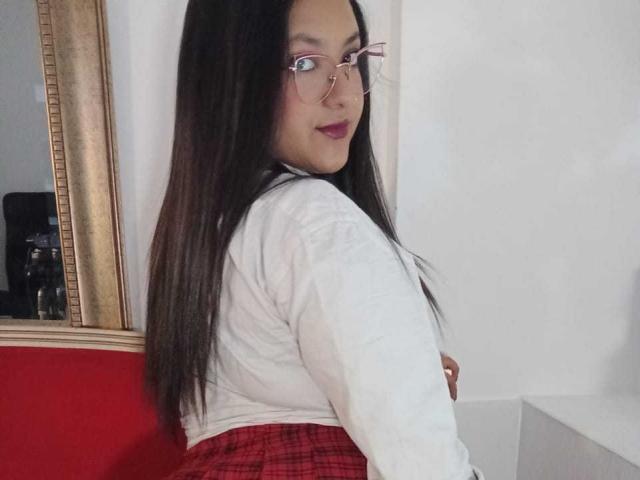 SafiMontiel with long black hair, glasses, white shirt, and red plaid skirt, looking over her shoulder, showing her ass. She's smiling with red lipstick.