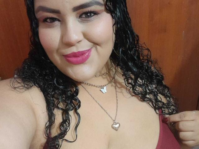 MelannieCarter with curly black hair, pink lipstick, and a heart necklace, smiling at the camera, showing off her tits with a hand on her red top.