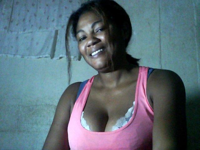 Jaiya, sorridente, indossa una canotta rosa, mostrando il seno con un reggiseno bianco che si intravede, pelle scura, capelli castani raccolti in una coda di cavallo, in piedi in una stanza poco illuminata..