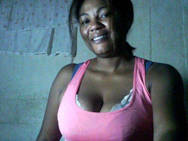 Jaiya, dark-skinned woman with medium breasts, pink tank top, white bra visible, smiling, brown hair in ponytail, standing, dimly lit, casual selfie.