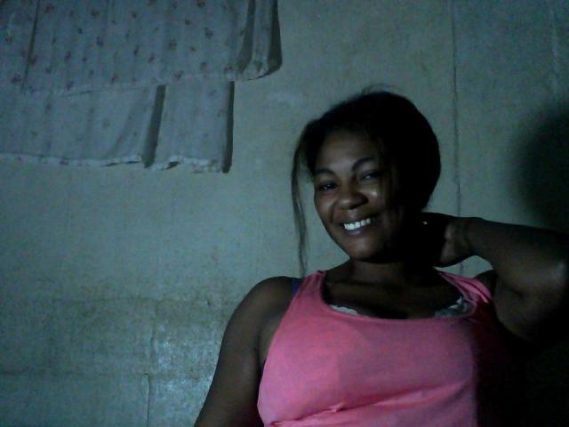 Jaiya with dark skin, smiling, wearing a pink tank top, showing her tits, arm raised, dimly lit, looking cute.