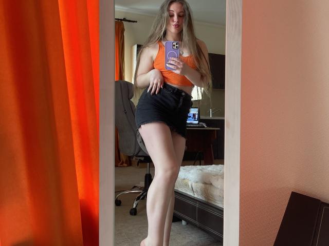 Mirror selfie of Dooris with long blonde hair, orange tank top, black high-waisted shorts, holding phone, standing, showing ass and legs, orange curtain on left, bed in background.