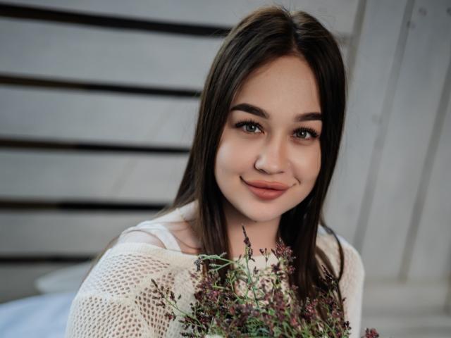 BettySweetty with long brown hair, green eyes, and a cute smile, holding a bunch of small purple flowers, wearing a white knit top, looking at the camera.