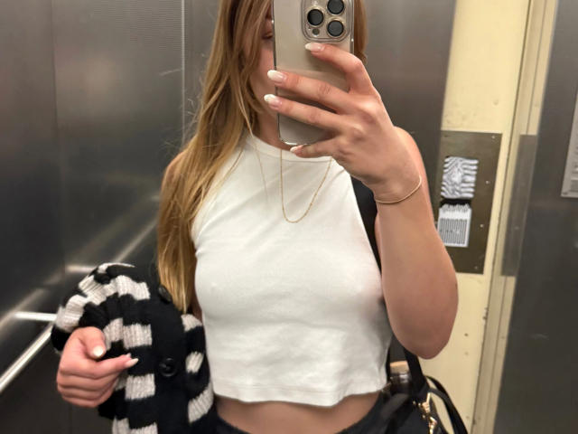 lanasativa-con cabello rubio largo, top blanco sin mangas que deja ver sus pechos, sosteniendo un teléfono, vistiendo una camisa a rayas negras y blancas, collar dorado, en un ascensor, tomando una selfie en el espejo..