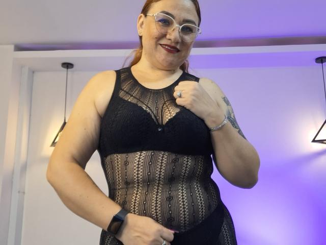 ElizabethMorrison, with glasses, black lace lingerie, and fishnet stockings, sits on a bed, showing her ass, and a tattoo on her chest, with a laptop and pillows.