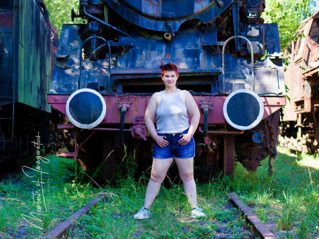 3Eine rothaarige Frau mit einem weißen Tanktop und blauen Shorts steht vor einem rostigen Zug, die Hände in die Hüften gestemmt, und zeigt ihr Gesäß, auf einem grasbewachsenen, sonnendurchfluteten Hof..
