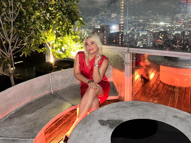 SophieVarto, una donna bionda con un seno di media grandezza, indossa un vestito rosso, seduta su un tetto curvo in cemento, con un pavimento in legno, luci della città sullo sfondo, pioggia sul vetro, sorridente..