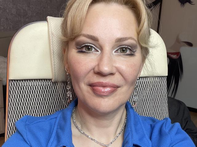NatalyFoxy69 heeft blond haar, groene ogen en ze glimlacht met roze lippen. Ze draagt een blauw shirt, een zilveren ketting en oorbellen, en zit in een stoel met een witte hoofdsteun..