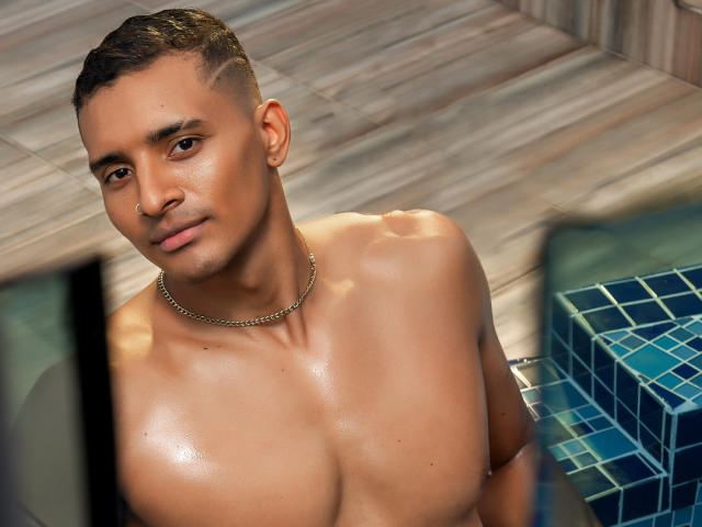 CarlosSmith, shirtless, with glistening brown skin, short braided hair, nose ring, chain necklace, and a subtle smile, standing by a tiled pool, looking up.