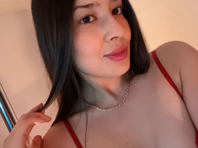 EmmaLombardi with long black hair, red bra straps, and a silver chain necklace, holding her hair, smiling with pink lips. Her tits are slightly visible.