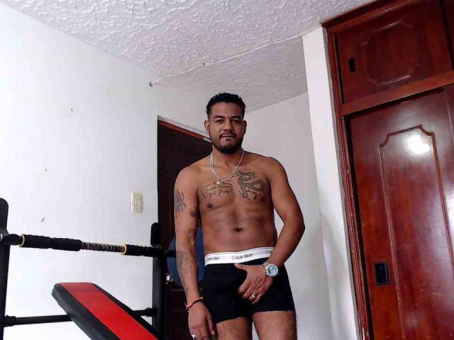 BladeFire, sem camisa, musculoso, tatuado, vestindo cuecas pretas da Calvin Klein, em pé em um quarto, mão direita em suas cuecas, armário de madeira à direita, equipamentos de ginástica à esquerda..