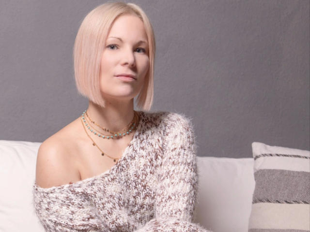 Sonnig-helles, warmes Licht, blonde Bob-Frisur, lockerer, flauschiger Pullover, Kette, sitzend auf einem weißen Sofa, Blick in die Kamera, leichtes Lächeln, weiches Licht, grauer Hintergrund..