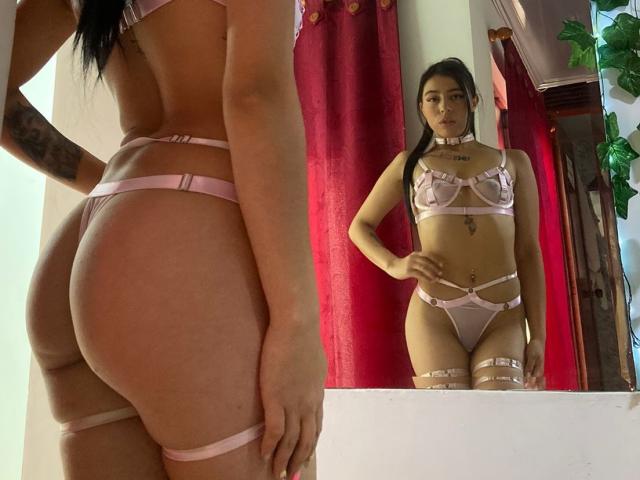 SaraHall in roze, open lingerie, die haar billen en borsten laat zien, staat voor een spiegel, waarbij haar vagina reflecteert, met een choker en tatoeages, in een kamer..