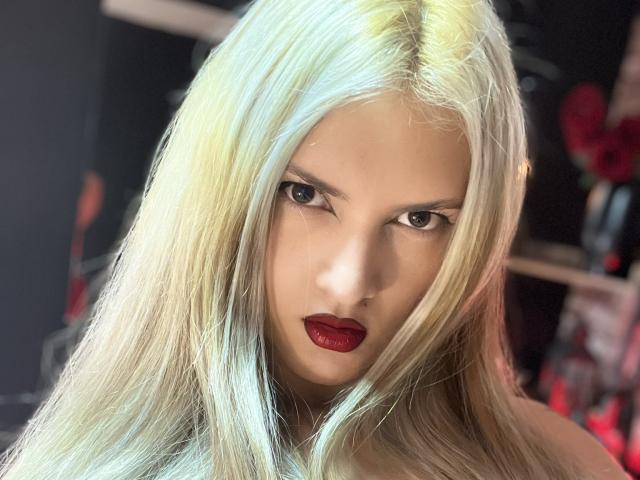 FiorellaNox with long, pale blonde hair, intense brown eyes, and dark red lipstick, staring directly at the camera, showing off a serious and sultry expression.