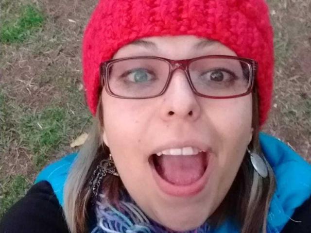 ArtemisFlyve porte un bonnet rouge en tricot, des lunettes rouges, et a la bouche grande ouverte, montrant ses dents. Elle a les yeux bleus, la peau claire, et une écharpe avec des motifs bleus et blancs..