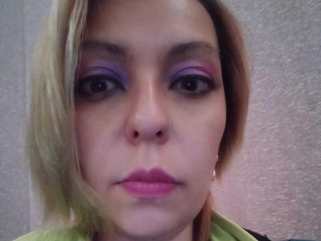 ArtemisFlyve heeft bleke huid, paarse oogschaduw, roze lippenstift en blond haar met een scheiding aan de zijkant. Ze draagt een groene blouse en heeft een neusring..