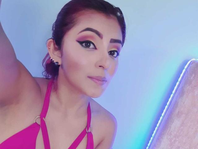 LizBonnet with dark brown hair in a ponytail, wearing a pink strappy bikini top, has thick black eyebrows, eyeliner, and a slight smile, showing her tits.