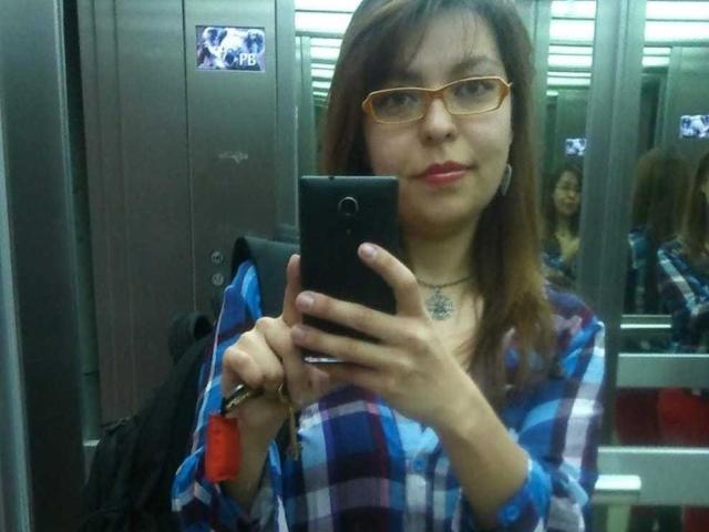 ArtemisFlyve, com cabelo castanho, óculos e batom vermelho, vestindo uma camisa azul xadrez, tirando uma selfie no espelho em um vestiário, segurando um celular preto e um chaveiro vermelho..