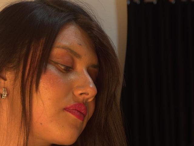 ChloeBeaumonth's face, eyes closed, brown hair, red lipstick, subtle freckles, eyeliner, sparkly eyeshadow, small star tattoo above left eyebrow, wearing elegant earrings, soft orange light.