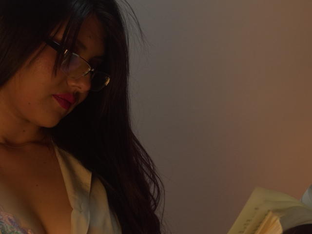ChloeBeaumonth with dark hair, glasses, and red lipstick, wearing an open shirt revealing her tits, looking down at a book. Dim lighting, soft shadows.