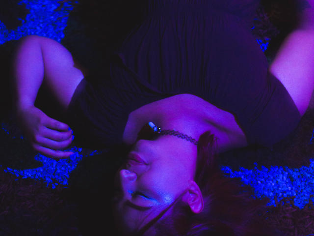 BeckyHuston acostada boca arriba, con luces moradas y azules, vestida con un vestido negro, un collar y sombra de ojos azul, con los brazos por encima de la cabeza, con una expresión relajada..