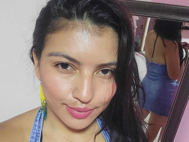 TamaraOriz ha la pelle olivastra, le labbra rosa, gli occhi neri e i capelli lunghi e scuri. Indossa degli orecchini giallo-verde e un top blu a fascia, mostrando la sua schiena in un riflesso allo specchio..