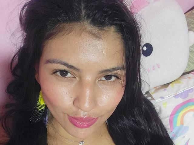TamaraOriz has a cum drip on her forehead, black wavy hair, pink lipstick, yellow earrings, and a cute unicorn pillow. Her face is glistening and she's smiling slightly.