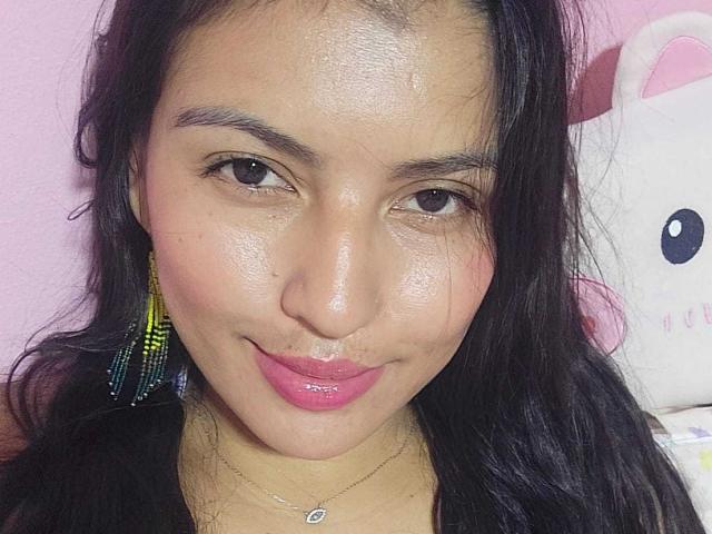 TamaraOriz hat ein glattes Gesicht mit rosafarbenem Lippenstift, braunen Augen und langen, schwarzen Haaren. Sie trägt bunte Ohrringe und eine Halskette mit einem kleinen Herzanhänger..