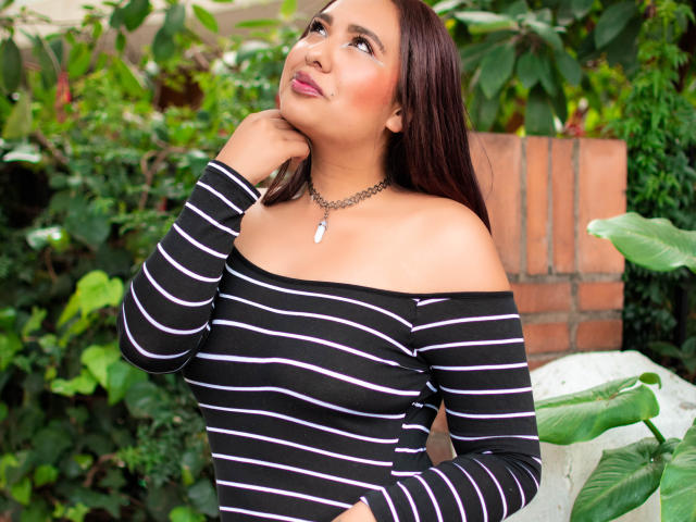 BeckyHuston with dark brown hair, wearing a black and pink striped off-shoulder top, looking up with a slight smile, hand near her neck, green leafy background.
