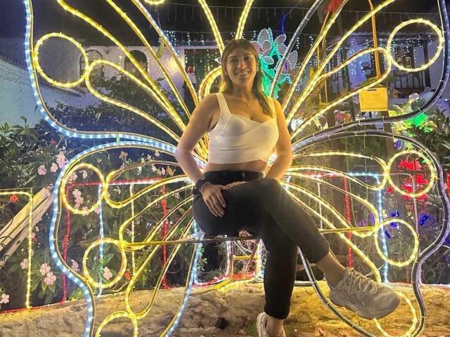 LunaBeya with tits in a white crop top, black pants, and white sneakers, sitting in front of a neon butterfly light sculpture, smiling at the camera.