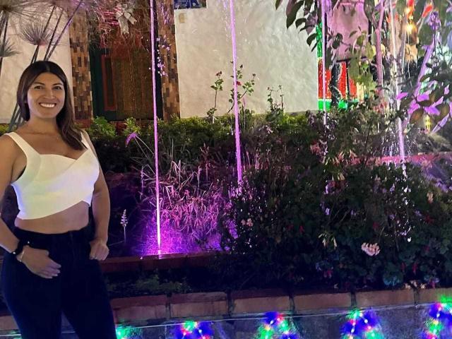 LunaBeya stands smiling with her tits barely covered by a white, cut-out crop top, wearing black pants. She's in front of a colorful, lit-up garden water feature.