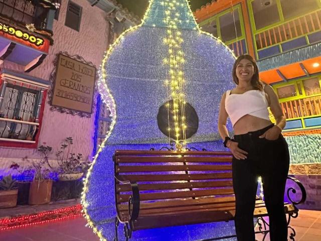 LunaBeya with tits covered by white crop top, black pants, standing next to wooden bench with blue and yellow lights, smiling at camera, night time, festive decorations.