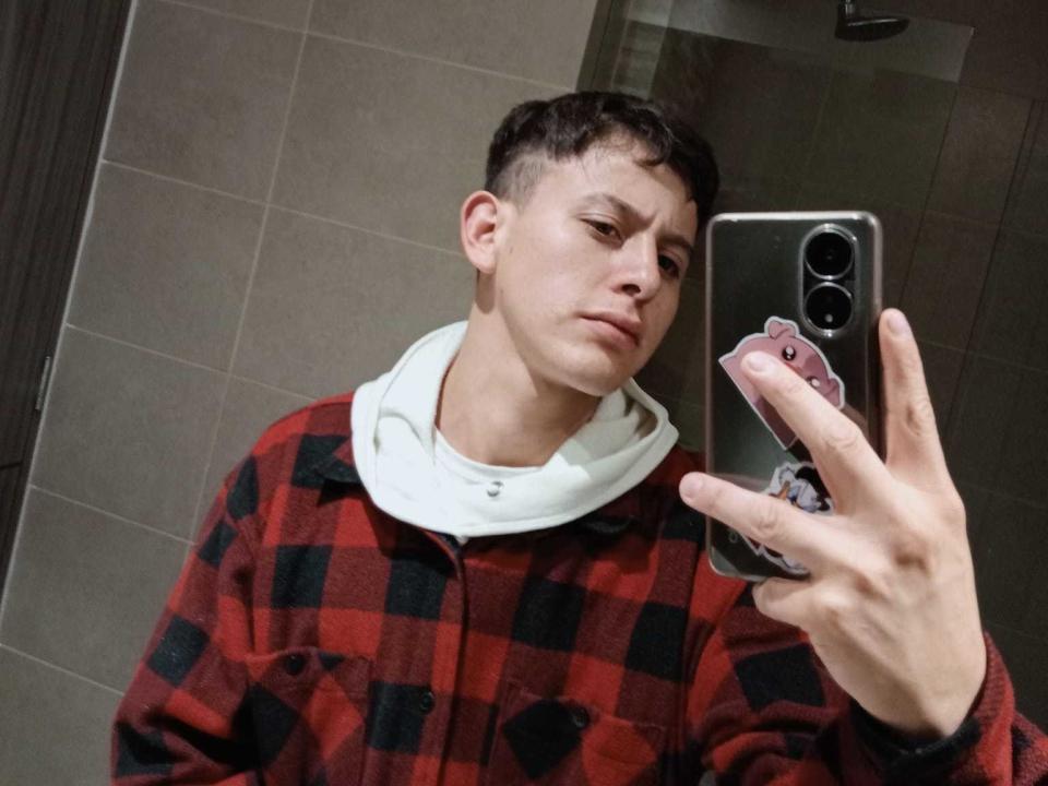 Alan Corss, con capelli castano corti, indossa una camicia a quadri rossa e nera, una felpa bianca e tiene il telefono per un selfie allo specchio, appare contrariato. Sullo sfondo si vede una parete piastrellata marrone. Il telefono ha un supporto rosa..