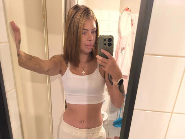 RoxyXXX - atraente, com cabelo castanho claro, top esportivo branco, calças brancas e um colar com uma cruz, tirando uma selfie no espelho, mostrando uma tatuagem no braço e um piercing no umbigo..