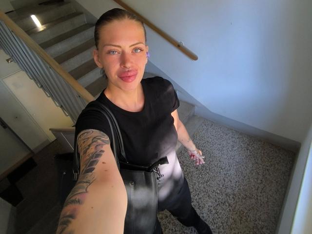 RoxyXXX, com tatuagens no braço direito, vestindo uma blusa preta, calças pretas e carregando uma bolsa preta, está tirando uma selfie no andar de baixo, fazendo beicinho com seus lábios rosados e olhando para a câmera..
