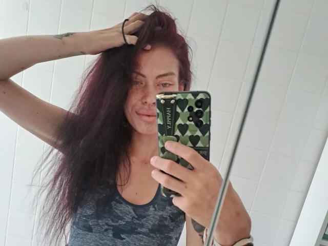 RoxyXXX – eine attraktive Frau mit zerzaustem, rotem Haar, die ein graues Tanktop mit Tarnmuster trägt und ein Handy mit einer grün-herzförmigen Hülle hält. Sie macht ein Selfie im Spiegel, wobei ihre Hand in ihrem Haar ist..