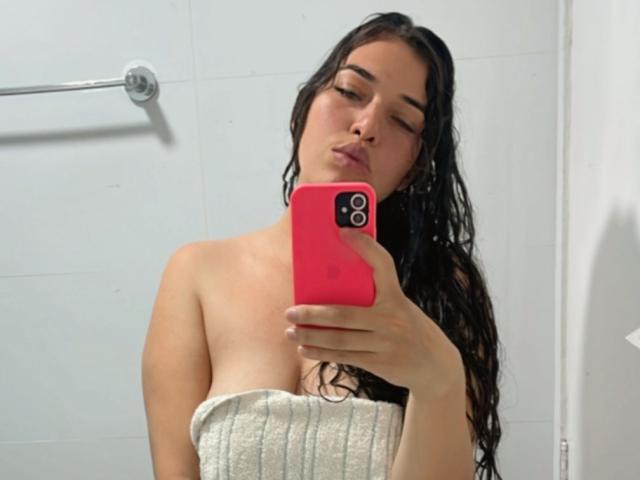 BelletinaXx, com seios grandes, vestindo uma blusa preta transparente com padrão geométrico e calças combinando, sentada em uma cama, mostrando tatuagens em seu braço direito e pulso esquerdo..