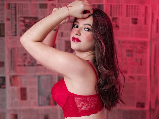 MadissonFontaine with long black hair, red lipstick, and a finger to her lips, showing a cute smile with a small mole on her cheek, topless.