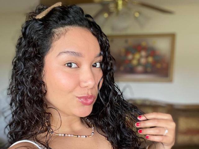 Ashlley69 met krullend zwart haar, bruine huid en rode lippenstift, die een ondeugende uitdrukking heeft, draagt een kralenketting en een ring, met rode nagels die haar haar aanraken..