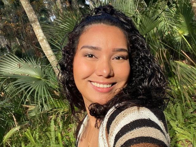 Ashlley69 has curly black hair, brown eyes, and a warm smile, wearing a striped shirt, standing in a sunny, green, leafy forest with palm fronds in the background.