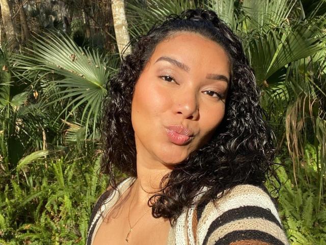 Ashlley69 with curly black hair, wearing a white top, beaded necklace, and red nails, pouting her glossy pink lips, and touching her hair with a cute, confident expression.