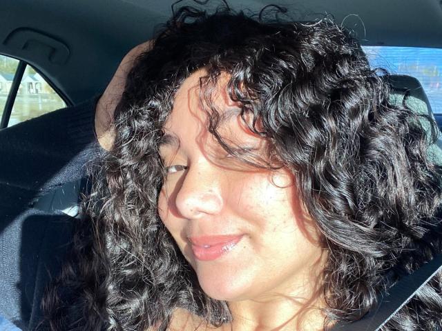 Ashlley69 a les cheveux noirs et bouclés, des yeux marrons, et elle a l'air boudeuse avec les lèvres légèrement entrouvertes. Elle porte un pull rayé, ce qui révèle sa peau bronzée et son petit nez rond..
