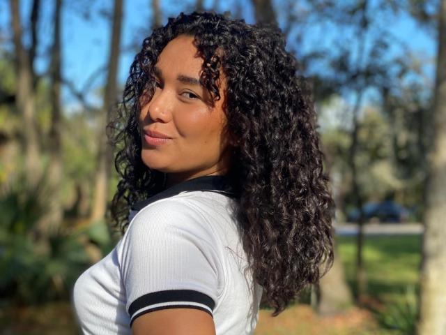 Ashlley69, con capelli scuri, pelle abbronzata e un leggero sorriso, indossa una giacca blu, tiene in mano degli occhiali da sole e si trova su una passerella di legno, guardando oltre la spalla verso la fotocamera..