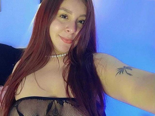 Ashlley69 con capelli neri ricci, pelle marrone media, indossa una camicia bianca con bordi neri, guarda oltre la spalla, sorriso leggero, luce solare sul viso, sfondo di foresta, cielo blu limpido..