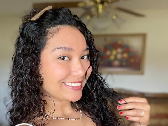 EmaJames tiene el pelo largo y castaño, piel clara, lápiz labial rojo y un pequeño tatuaje de una hoja en el hombro derecho. Está sin ropa, sonriendo a la cámara con la mano izquierda en el pelo..
