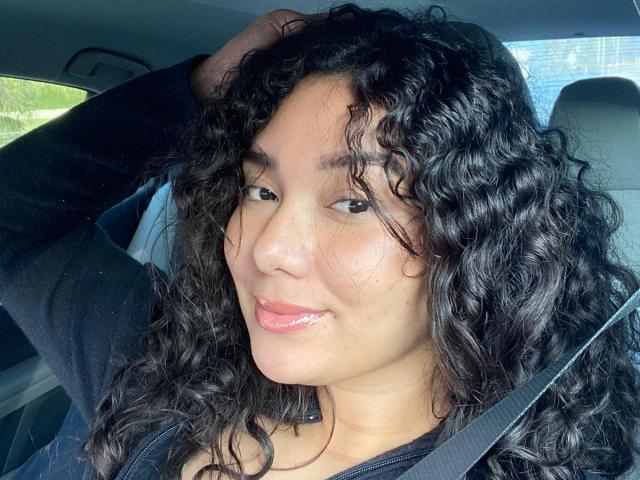 EmaJames, con i suoi lunghi capelli neri, il rossetto rosso e un leggero sorriso, mostra il seno indossando una canottiera nera a spalline sottili. Indossa degli auricolari verdi..