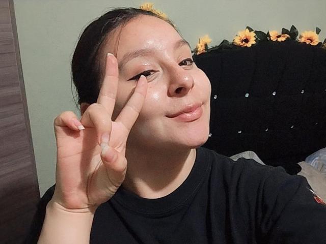 EmaJames, ein asiatisches Mädchen mit rotbraunen Haaren, streckt ihre rosa Zunge heraus, blickt nach oben, trägt Diamant-Ohrringe, im Hintergrund eine weiße Wand, ein süßes und verspieltes Gesicht..