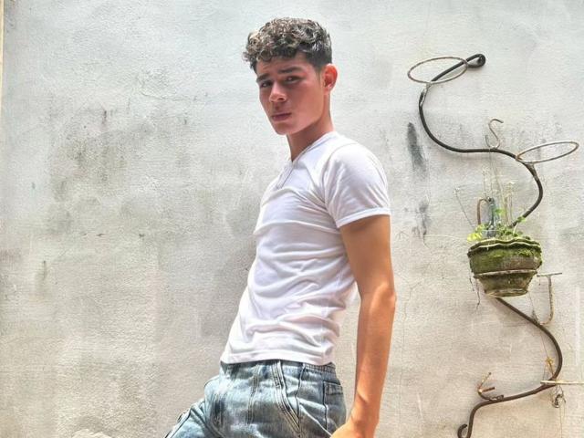 EmaJames, con i capelli rossi raccolti in una coda di cavallo, indossa una maglietta nera, orecchini a cerchio dorati, mostra un segno di pace, rivela dei denti metallici ed è sorridente. Le sue natiche non sono visibili..
