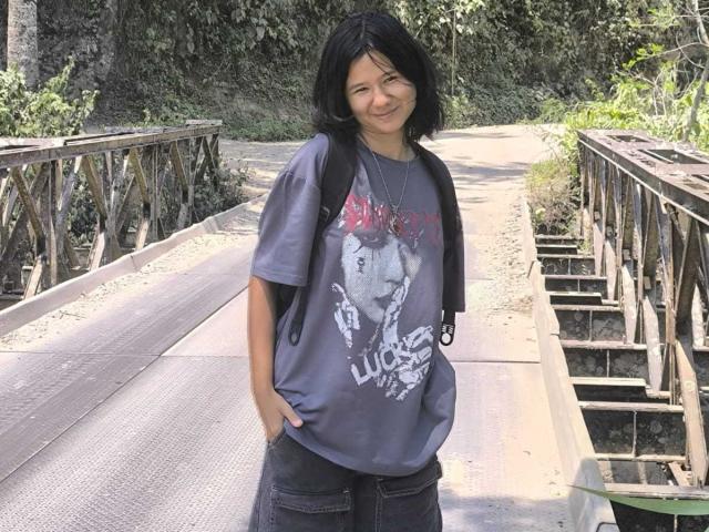 Sophie Smith, con cabello oscuro y ondulado, una camiseta negra y pantalones cortos coloridos, sentada en el césped, mirando hacia arriba con una ligera sonrisa, sosteniendo un palo para selfies en su mano derecha..